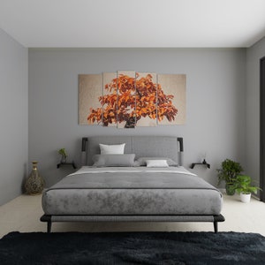 A modern bedroom featuring a gray upholstered bed with a matching comforter and pillows. Above the bed is a multi-panel artwork depicting an orange tree. The room includes a black rug, decorative vase, and potted plants.