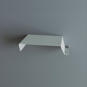 A minimalist, white metal shelf with a clean, modern design. The shelf has a flat top and a downward-sloping front edge, with a small, rectangular support bracket. The shelf is mounted on a gray wall.