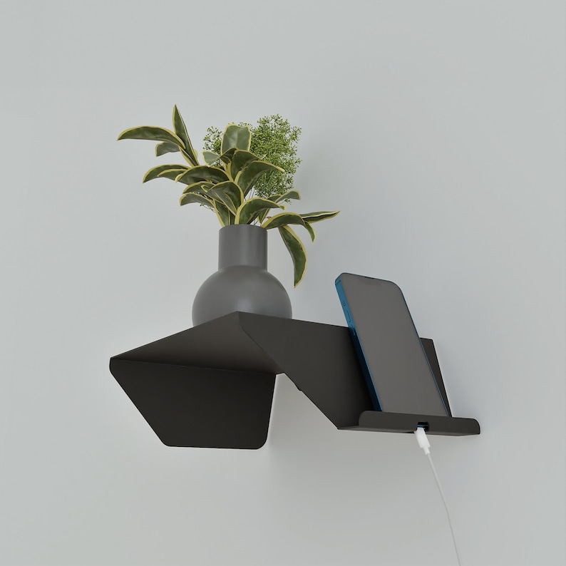 A modern black wall-mounted shelf with a built-in phone stand. A smartphone is charging on the stand. A gray vase with green plants sits on the shelf. The shelf is mounted on a white wall.