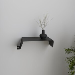 A black metal wall shelf with a unique, angular design. A small, dark vase holds delicate, dried branches. The shelf is mounted on a white wall, creating a minimalist aesthetic.