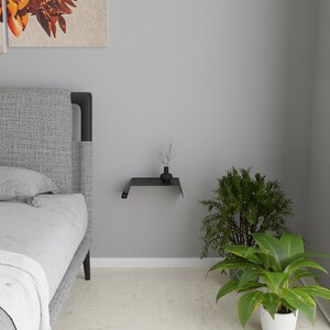 A black metal floating shelf mounted on a gray wall. A small black vase with dried plants sits on the shelf. A bed with a gray headboard is on the left, and green plants are on the right.