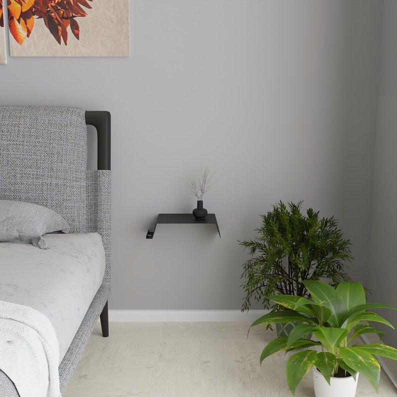 A black metal floating shelf mounted on a gray wall. A small black vase with dried plants sits on the shelf. A bed with a gray headboard is on the left, and green plants are on the right.