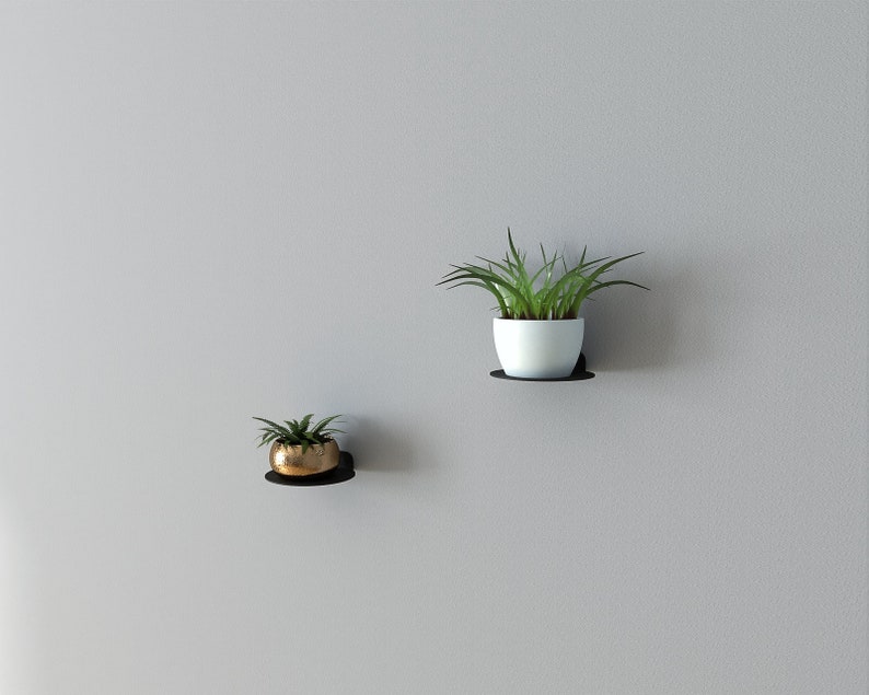 May include: Two black metal wall shelves with plants in pots. The top shelf has a white pot with a green plant. The bottom shelf has a gold pot with a green plant.