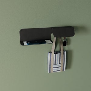 May include: A black metal wall shelf with a small, rectangular shelf and a hook. A blue and white striped tote bag hangs from the hook. A black phone and a black watch are on the shelf.