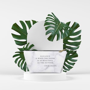 May include: A white marble-patterned cosmetic bag with a black zipper and gold-colored pull. The bag features the quote "Elegance is when the inside is as beautiful as the outside - Coco Chanel." Large green monstera leaves surround the bag.