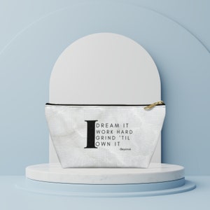 May include: White canvas cosmetic bag with a black zipper and gold-colored pull. The bag features the quote "I DREAM IT WORK HARD GRIND 'TIL OWN IT" in black text, attributed to Beyoncé.