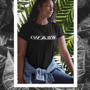 以下が含まれることがあります： 白い文字で「I.W.A.B.W.」と書かれた黒いTシャツ、デニムジャケットとジーンズを着用。背景には緑のシダの葉があります。画像は、グレースケールの葉を背景に白い枠で囲まれています。
