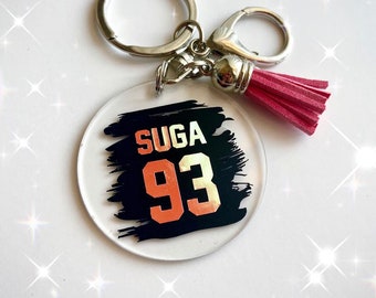 May include: A round clear acrylic keychain with a black and white brushstroke design. The design features the text "SUGA 93" in gold and orange lettering.