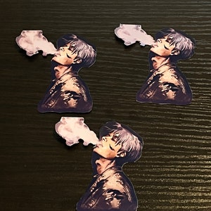 May include: Three identical stickers of a man with short, dark hair, wearing a brown jacket, blowing out a puff of smoke. The man is looking to the right. The stickers are on a dark wood surface.