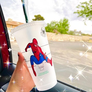 Könnte beinhalten: Ein klarer Plastikbecher für kalte Getränke mit einem Spider-Man-Design in Rot, Blau und Schwarz. Der Becher hat einen Deckel, einen grünen Strohhalm und ein schwarzes Spinnenlogo. Der Becher wird vor einem verschwommenen Hintergrund im Freien gehalten.