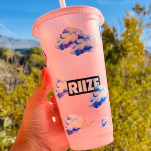 Puede incluir: Un vaso reutilizable rosa con una pajita, con un diseño de nubes y la palabra "RIIZE" en letras negras.