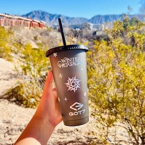 Pode incluir: Um copo cinza escuro com tampa e canudo pretos. O copo apresenta o texto "WINTER HEPTAGON" e "GOT7" com desenhos de flocos de neve e estrelas. O fundo mostra uma vista panorâmica de montanhas e folhagem.