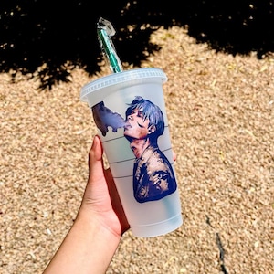 Puede incluir: Un vaso reutilizable de plástico transparente con una pajita verde. El vaso tiene un diseño impreso de una persona con cabello azul y una chaqueta negra. La persona está fumando un cigarrillo.