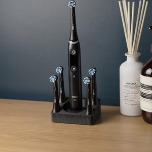 Könnte beinhalten: Eine schwarze elektrische Zahnbürste mit mehreren Bürstenköpfen, präsentiert in einem schwarzen Halter. Die Zahnbürste und die Köpfe haben blaue und weiße Borsten. Eine weiße Vase mit Schilf und eine dunkelbraune Flasche sind ebenfalls zu sehen.
