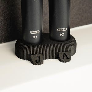 Puede incluir: Soporte negro para cepillos de dientes con dos cepillos de dientes eléctricos Oral-B iO. El soporte tiene un diseño acanalado y presenta las letras "J" y "A" en la parte delantera. Los cepillos de dientes son negros.