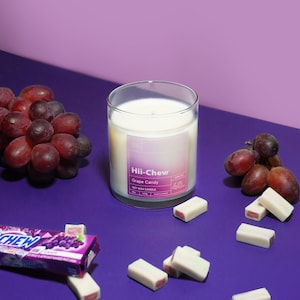 Puede incluir: Un tarro de vela de vidrio con una vela de cera de soja blanca en su interior. La etiqueta dice "Hii-Chew Grape Candy Soy Wax Candle 4oz". La vela est&aacute; rodeada de uvas moradas y cuadrados de caramelo blanco y rosa sobre una superficie morada.