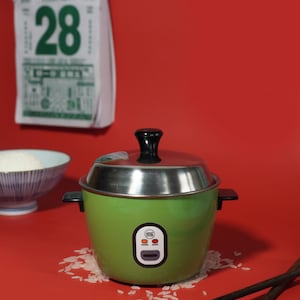 May include: A green rice cooker with a silver lid and black handle. The rice cooker has a digital display with the words "Warm" and "Cook". There are grains of rice scattered around the rice cooker and two wooden chopsticks.