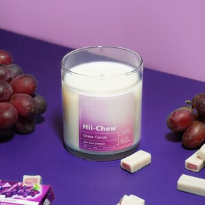 Puede incluir: Un tarro de vidrio para velas con una etiqueta degradada en morado y rosa que dice "Hii-Chew Grape Candy Soy Wax Candle 40h Burn Time 227g Made in Canada". La vela est&aacute; rodeada de uvas moradas y barras de caramelo blancas y rosadas sobre una superficie morada.
