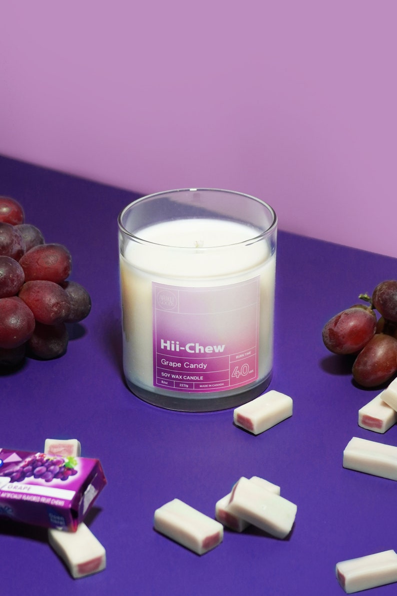 Puede incluir: Un tarro de vidrio para velas con una etiqueta degradada en morado y rosa que dice "Hii-Chew Grape Candy Soy Wax Candle 40h Burn Time 227g Made in Canada". La vela est&aacute; rodeada de uvas moradas y barras de caramelo blancas y rosadas sobre una superficie morada.
