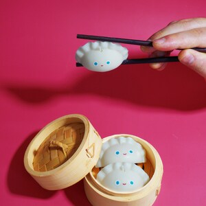 May include: Two white dumpling-shaped candles with blue eyes and a smile are inside a wooden steamer basket. A third dumpling candle is being held by chopsticks.