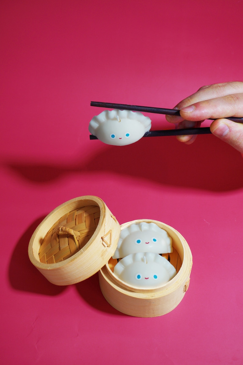 May include: Two white dumpling-shaped candles with blue eyes and a smile are inside a wooden steamer basket. A third dumpling candle is being held by chopsticks.