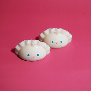 May include: Two white candles shaped like dumplings with blue eyes and a red smile. The candles are on a pink background.