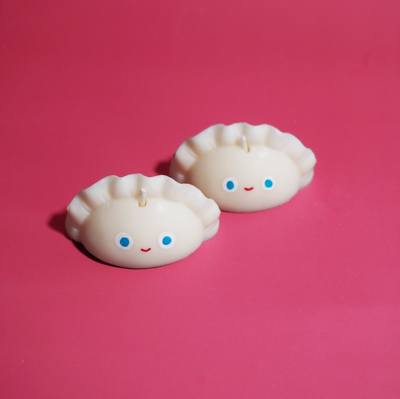 May include: Two white candles shaped like dumplings with blue eyes and a red smile. The candles are on a pink background.