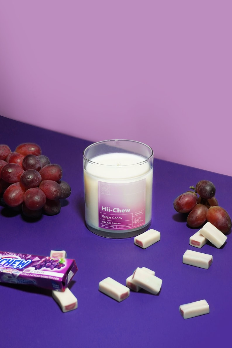 Puede incluir: Un tarro de vela de vidrio con una vela de cera de soja blanca en su interior. La etiqueta dice "Hii-Chew Grape Candy Soy Wax Candle 4oz". La vela est&aacute; rodeada de uvas moradas y cuadrados de caramelo blanco y rosa sobre una superficie morada.