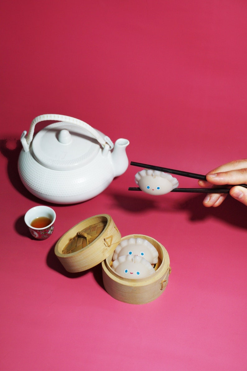 May include: Two white dumplings with blue eyes and a smiling mouth are in a bamboo steamer. A white teapot with a brown handle is on a pink surface. A hand is holding a pair of chopsticks with a dumpling on them.