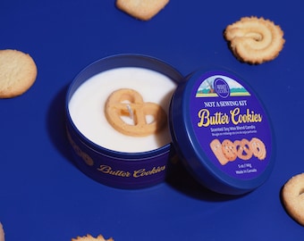 May include: A blue tin candle with a pretzel-shaped wax design, labeled "Butter Cookies." The candle is surrounded by various butter cookies on a blue surface. The label reads "NOT A SEWING KIT" and "Scented Soy Wax Blend Candle."