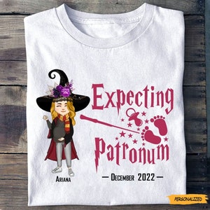 May include: White t-shirt with a pink graphic of a pregnant witch wearing a black hat and a red cape. The graphic says "Expecting Patronum - December 2022" and includes a pacifier, baby footprints, and stars. The witch's name is "Ariana".