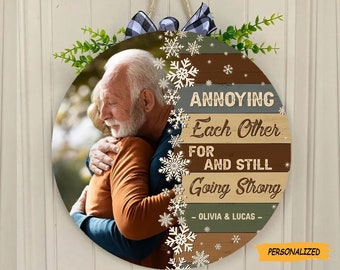 May include: A round wooden sign with a photo of an older couple hugging. The sign has a rustic woodgrain background with a gray, brown, and green striped pattern. The text on the sign reads "Annoying Each Other For And Still Going Strong - Olivia & Lucas - Personalized".