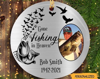 May include: A silver Christmas ornament with a black silhouette of a fish and birds flying upwards. The ornament has the text "Gone Fishing in Heaven" and a photo of a man holding a fish. The text "Bob Smith 1942-2021" is also on the ornament.