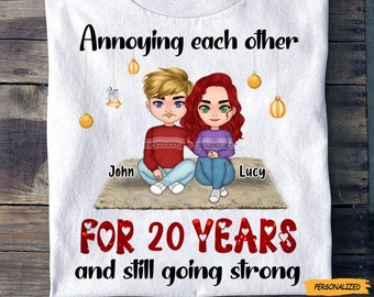 May include: A white t-shirt with a Christmas-themed design featuring a cartoon couple, John and Lucy, sitting together. The text reads "Annoying each other for 20 years and still going strong".
