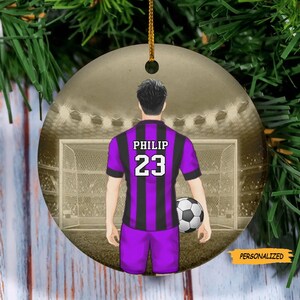 May include: A purple and black soccer jersey with the name "PHILIP" and the number "23" on the back. The jersey is worn by a player standing in front of a soccer goal. The player is holding a soccer ball.