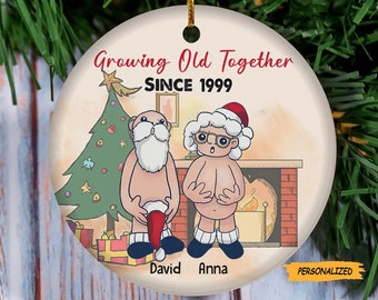 May include: A white ceramic Christmas bauble featuring a cartoon illustration of a couple dressed as Santa Claus. The bauble has the text "Growing Old Together Since 1999" and the names "David" and "Anna".