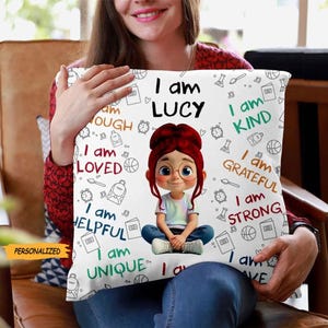 May include: Square white cushion featuring a cartoon girl with red hair and glasses. The cushion has the text "I am LUCY" and positive affirmations such as "I am KIND" and "I am STRONG". The word "PERSONALIZED" is also visible.