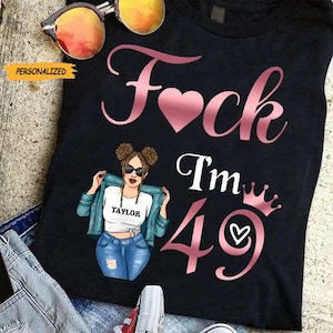 May include: Black t-shirt with the text "F*ck I'm 49" in pink script and a heart. Features a cartoon illustration of a woman wearing sunglasses, a white shirt with the name "Taylor", a blue jacket, and jeans. Sunglasses are on top.