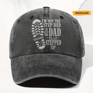 May include: A charcoal gray baseball cap featuring an embroidered white graphic of a boot print and the text: I'M NOT THE STEP DAD I'M JUST THE DAD THAT STEPPED UP HENRY. A small orange rectangular tag in the upper right corner reads PERSONALIZED.