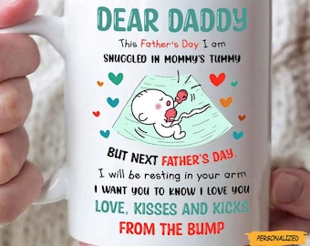 May include: White ceramic mug with the text "DEAR DADDY" in teal. The mug features a cartoon ultrasound image with a baby and the text "This Father's Day I am snuggled in Mommy's tummy". The mug also has the text "But next Father's Day, I will be resting in your arm".