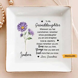 May include: White ceramic trinket dish with a gold rim. The dish features a purple floral design and a message that reads "To My Granddaughter Whenever you feel overwhelmed, remember whose granddaughter you are and straighten YOUR CROWN. Always remember you are Braver than you think, Stronger than you seen and Loved more than you know. Love, Grandma". The dish is personalized with the name "Isadora".