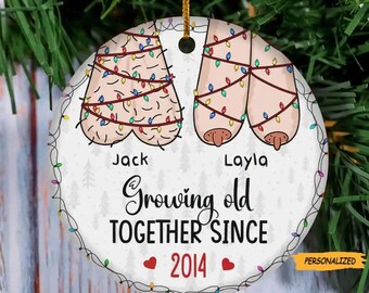 May include: White round Christmas bauble featuring a cartoon illustration of two figures adorned with Christmas lights. The text reads "Jack & Layla Growing Old Together Since 2014".