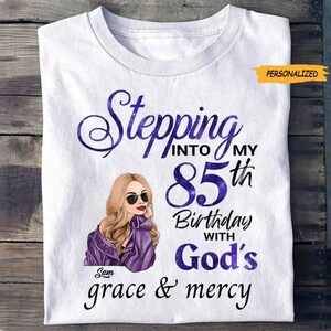 May include: White t-shirt with a graphic design. The design features the text "Stepping into my 85th Birthday with God's grace & mercy" in purple script. An illustration of a woman wearing sunglasses and a purple jacket is also included.
