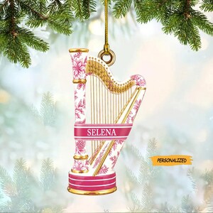 May include: A personalized harp-shaped Christmas ornament. The ornament features a white background with pink floral patterns and gold accents. The name "SELINA" is displayed on a pink banner. The word "PERSONALIZED" is also visible.
