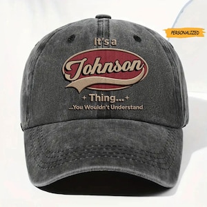 May include: A charcoal gray baseball cap with a distressed finish. The front features a maroon oval emblem with the text "It's a Johnson Thing... You Wouldn't Understand" in cream and brown lettering. A small orange "PERSONALIZED" tag is visible.
