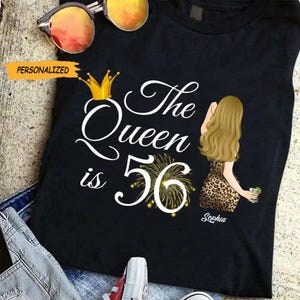 Peut inclure: T-shirt noir avec l'inscription "The Queen is 56" et un dessin d'une femme en robe léopard. Le t-shirt présente également une couronne dorée et un motif de feux d'artifice. Le mot "Personalized" est dans une bannière jaune.