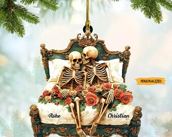May include: A decorative ornament featuring two skeleton figures in a bed, surrounded by red roses. The bed has a teal and gold frame. The names "Ashe" and "Christian" are printed on the bed. The word "PERSONALIZED" is also printed on the ornament.
