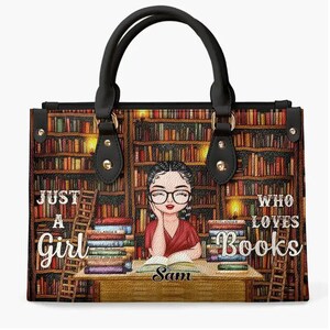 Puede incluir: Un bolso tote de cuero negro con una ilustraci&oacute;n colorida de una ni&ntilde;a leyendo un libro en una biblioteca. El bolso tiene una cremallera dorada y herrajes dorados. El texto "Just a Girl Who Loves Books" est&aacute; impreso en el bolso. El nombre "Sam" est&aacute; impreso en la parte inferior del bolso.