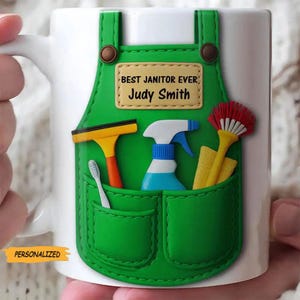May include: White ceramic mug featuring a green apron design with cleaning tools in the pockets. The apron has a name tag that reads "BEST JANITOR EVER" and "Judy Smith". The mug also has the word "PERSONALIZED" on the bottom.