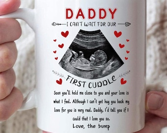 May include: White ceramic mug with the text "DADDY I CAN'T WAIT FOR OUR FIRST CUDDLE" and a sonogram image. Red heart graphics are scattered around the image. The mug is held by a person.